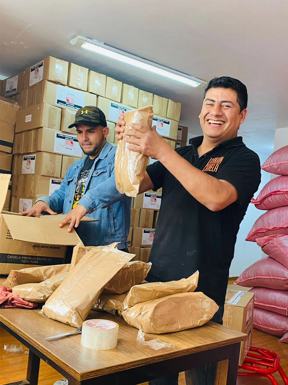 A smiling man in a black "Lankan Canela" polo shirt holds up a bag of cinnamon powder in a warehouse setting, while a colleague in a denim jacket and camouflage cap assembles a cardboard box behind him. The background is filled with tall stacks of labeled shipping cartons and large pink sacks, highlighting the busy packaging and distribution operations.