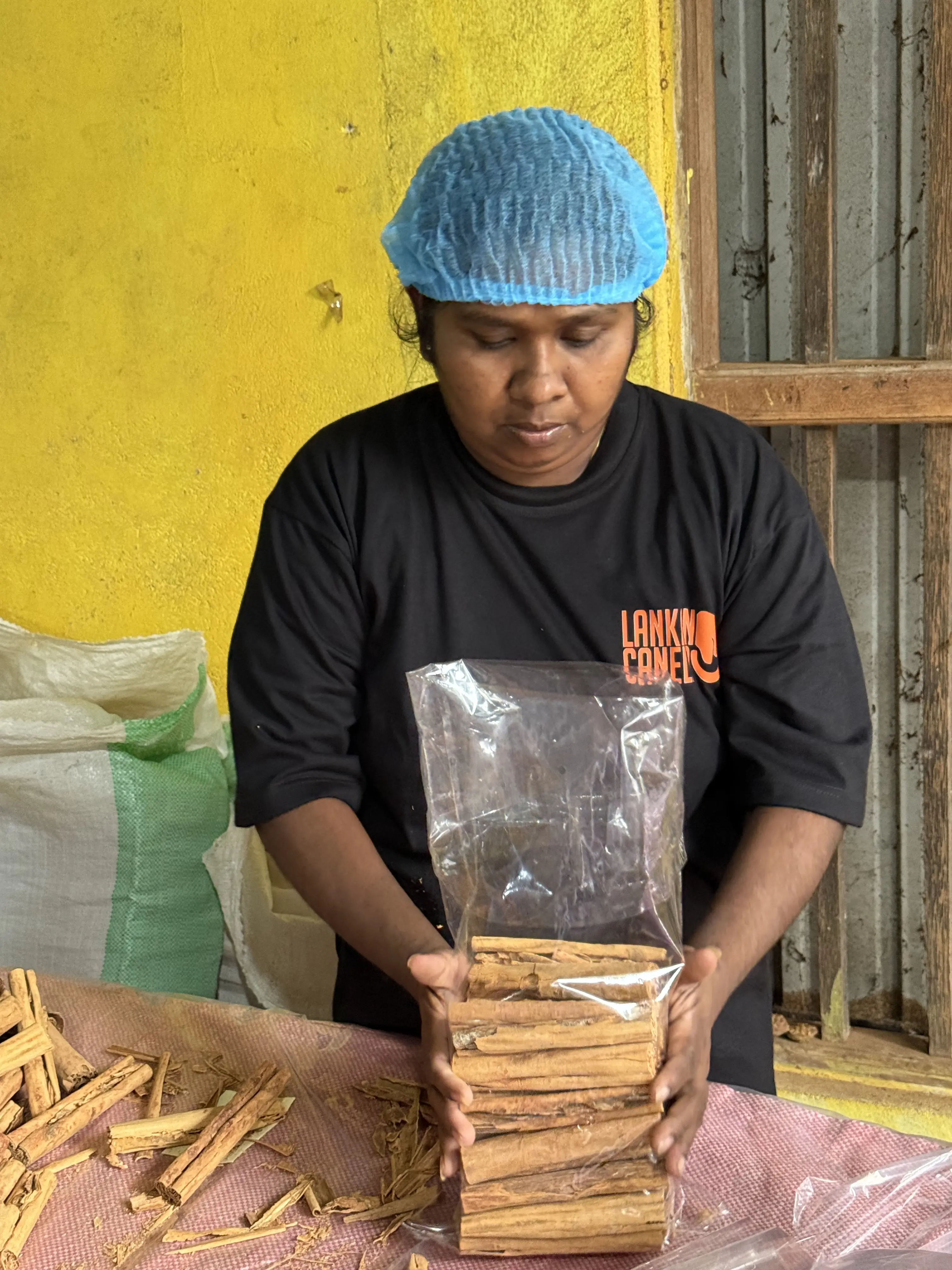A female worker, wearing a blue hairnet and a black T-shirt featuring the orange "Lankan Canela" logo, holds a clear plastic package filled with cinnamon quills, inspecting the product quality before final packing.