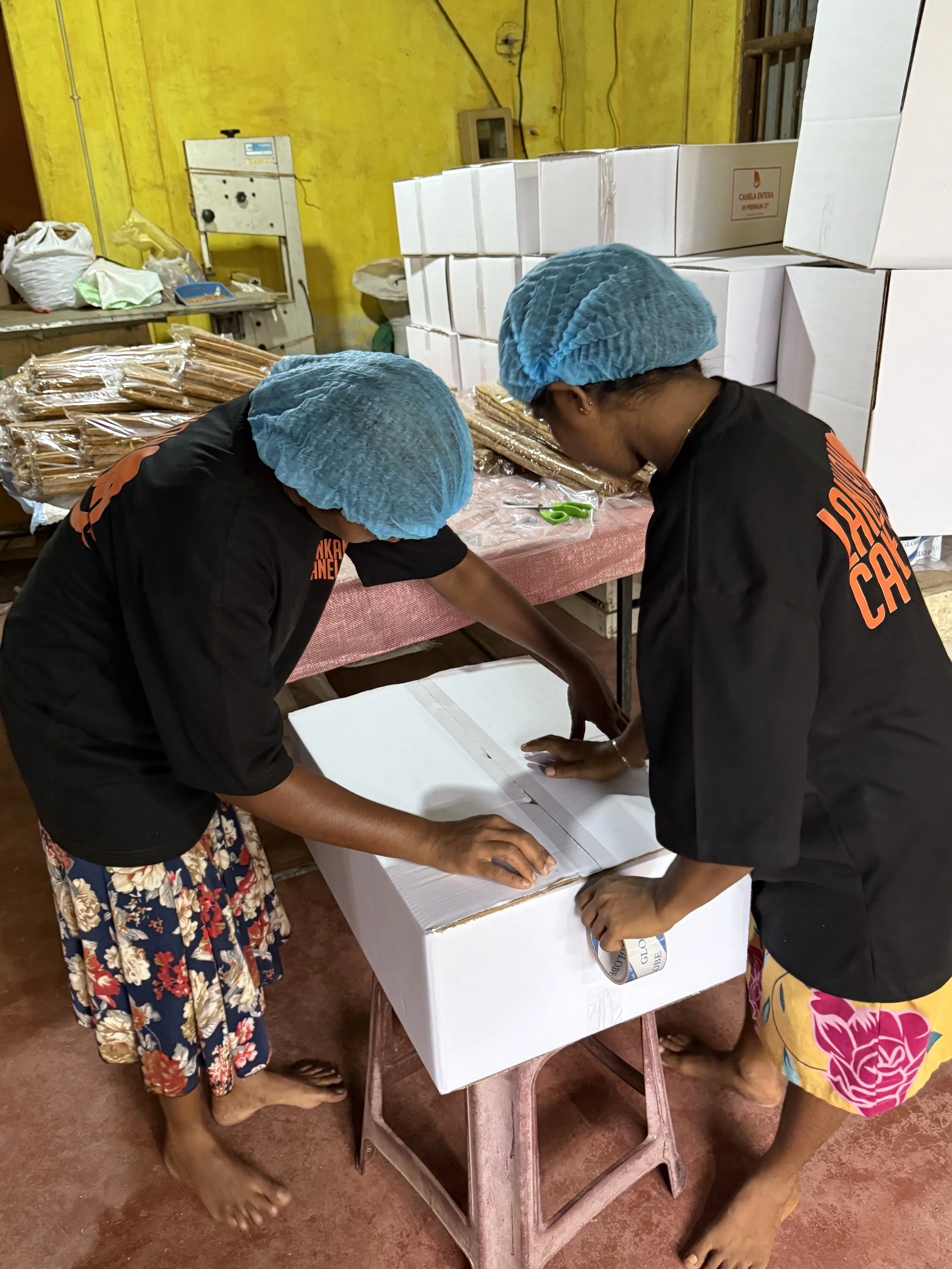 Two female workers, dressed in blue hairnets and black "Lankan Canela" branded T-shirts, work together to seal a white shipping carton with packing tape. The scene takes place in a processing facility with stacks of finished boxes and work tables holding cinnamon bundles visible in the background.