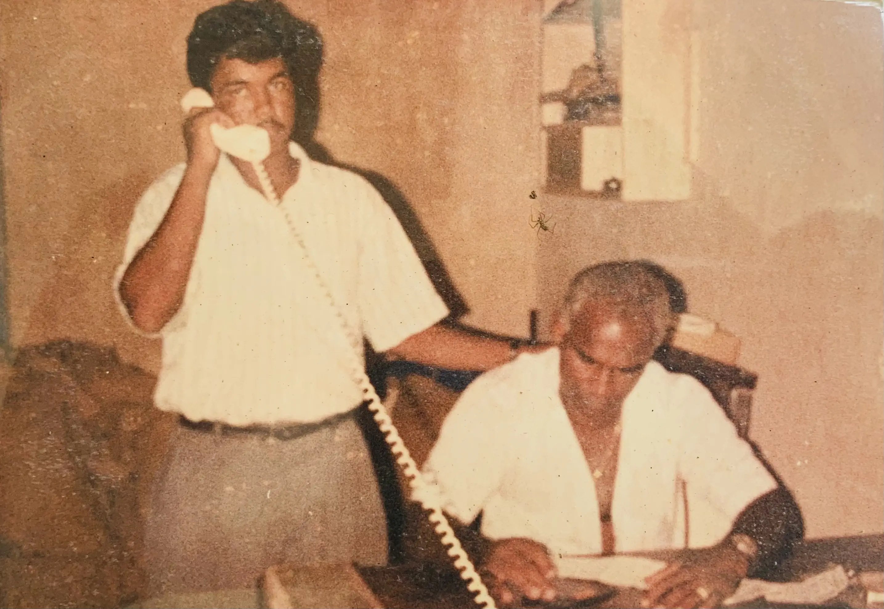Vintage, slightly faded photograph of two men in an office setting—one standing and speaking on a corded landline telephone, the other seated at a desk writing in a ledger—capturing an early-era business workspace.