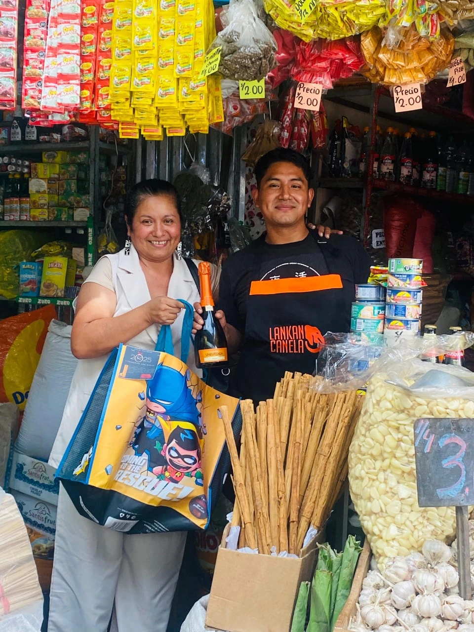 A man wearing a black apron with the orange "Lankan Canela" logo poses with a smiling female customer in a bustling market stall. The woman holds a bottle of sparkling wine and a reusable shopping bag, while a box of long cinnamon quills sits prominently in the foreground next to a large bag of peeled garlic and under rows of hanging spice packets.