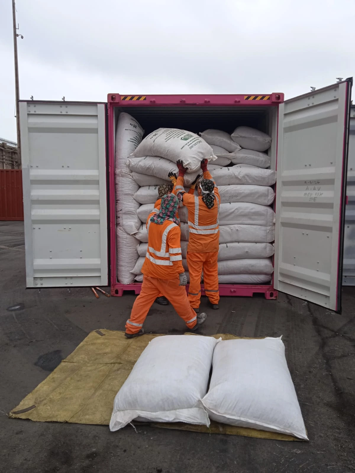 Three workers wearing bright orange safety jumpsuits and patterned head coverings work together to stack large white sacks inside a pink shipping container. The container is packed floor-to-ceiling with the bulk bags, while two additional sacks rest on a yellow tarp in the foreground, ready for loading.